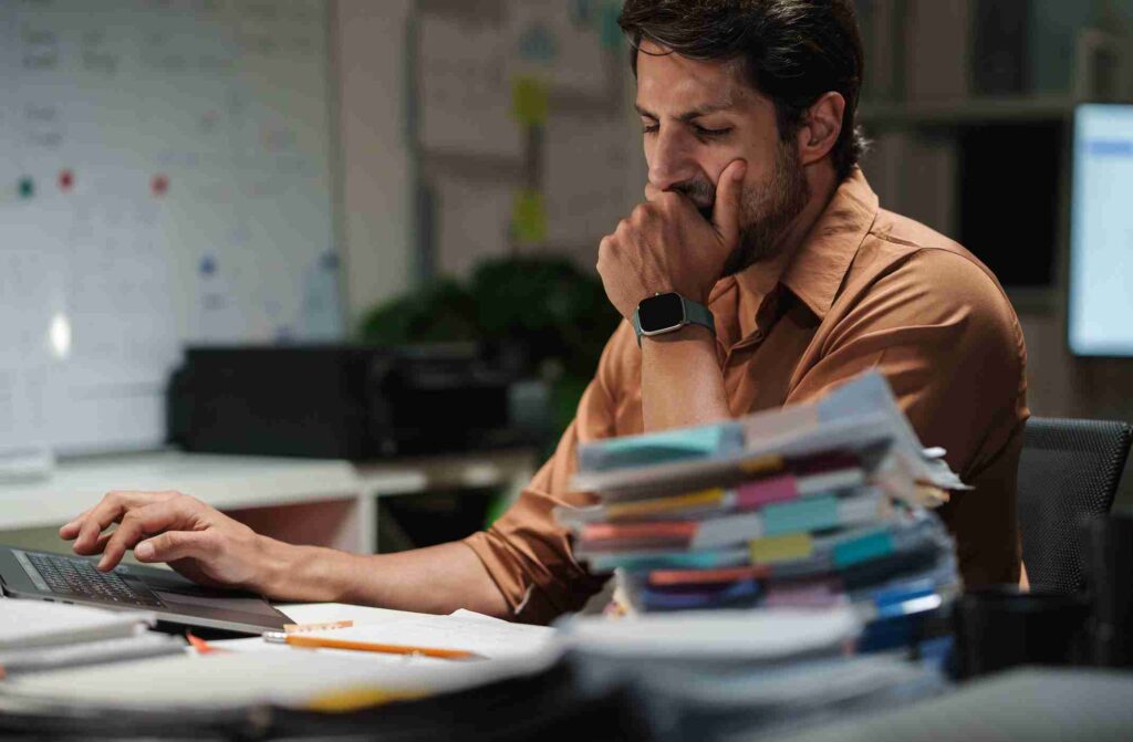 A man sitting at his desk resting his chin on his hand next to a tall stack of papers.